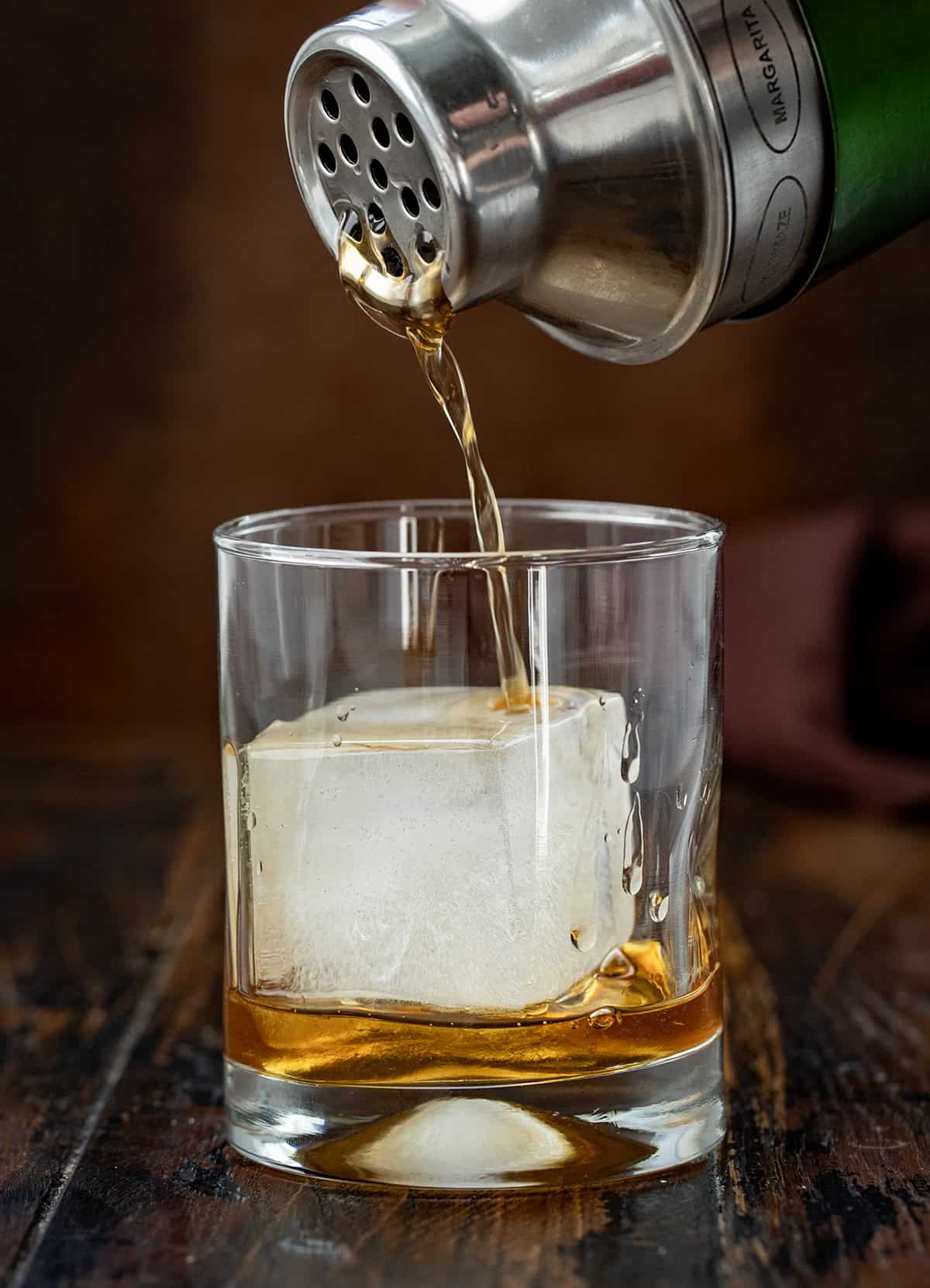 Warm, cozy, and perfectly spiced for fall, this Maple Old Fashioned is a classic cocktail with a seasonal twist! Whether you’re winding down after a busy day or serving a cocktail at a cool-weather gathering, this drink is easy to make and sure to impress! Pouring Maple Old Fashioned into a glass with a square ice cube.