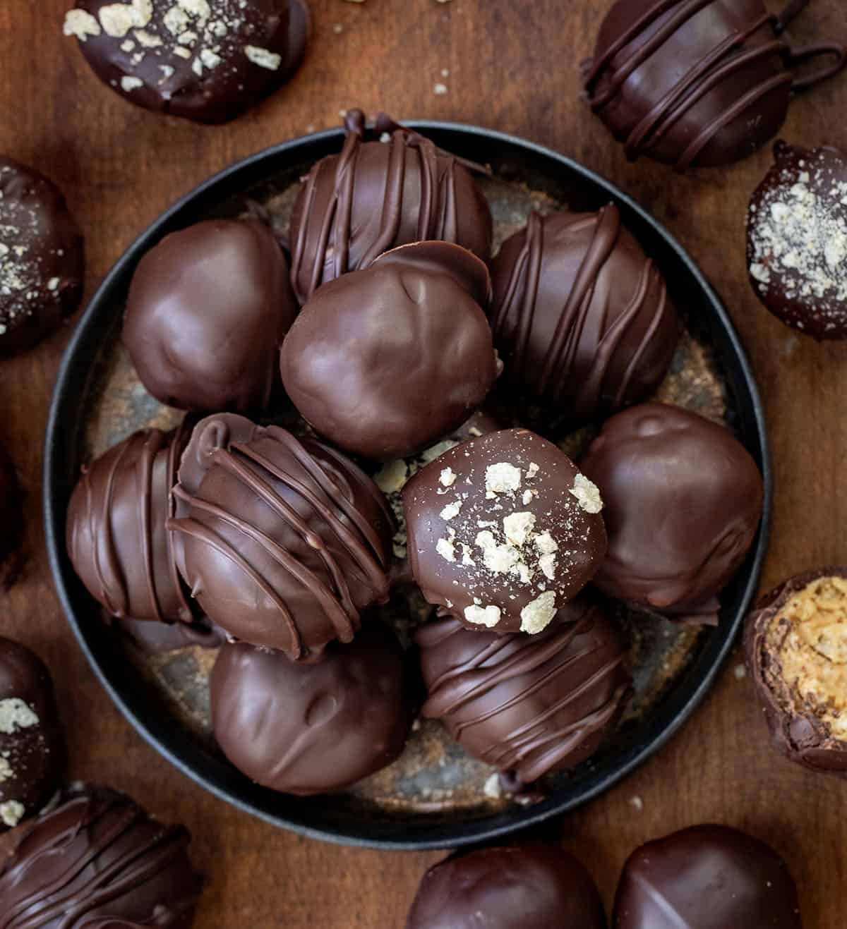 These Peanut Butter Balls with Rice Krispies are sweet, crunchy, and totally irresistible! Creamy peanut butter meets crispy cereal and a rich chocolate coating, making them the perfect treat! Small black platter of Peanut Butter Balls with Rice Krispies on wooden table from overhead.