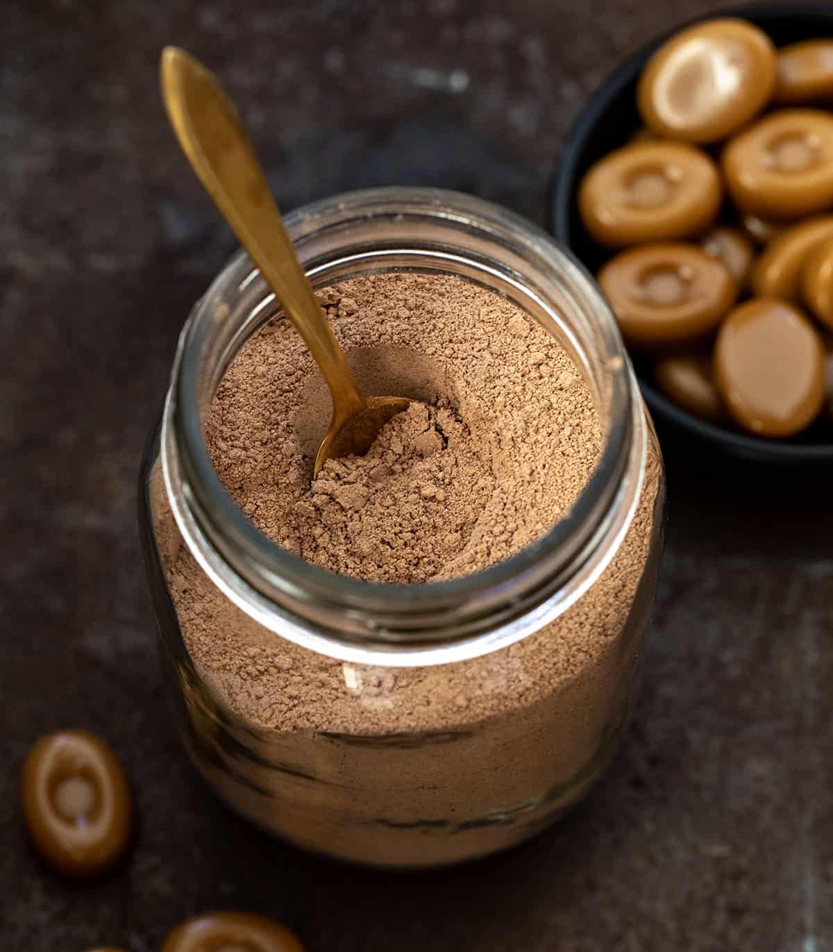 Glass mason jar filled with Salted Caramel Hot Cocoa Mix on a wooden table surrounded by hard caramels.