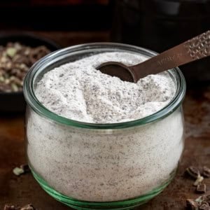 Glass container of Andes Hot Cocoa Mix with a measuring spoon in it on a dark table.