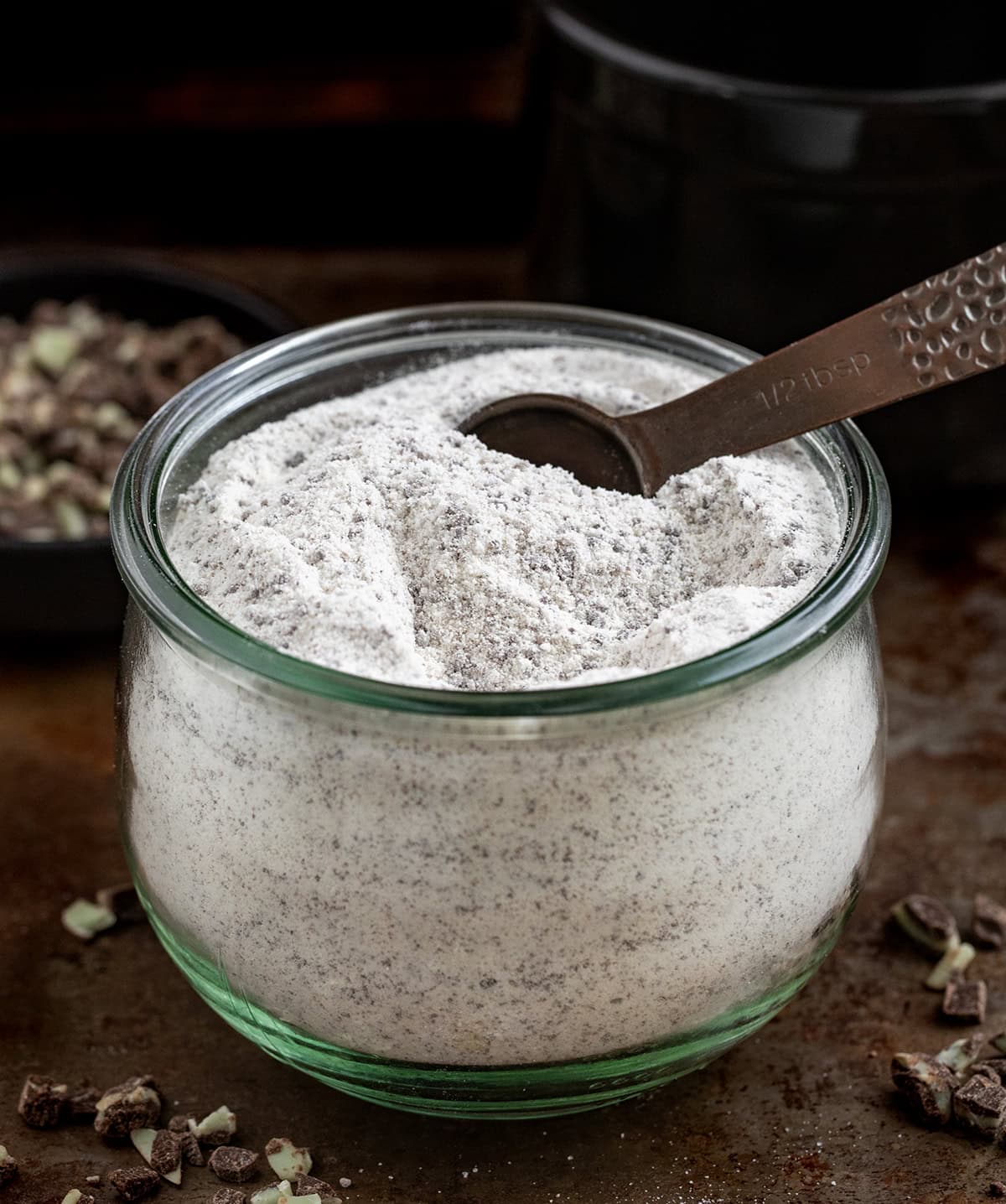 Glass container of Andes Hot Cocoa Mix with a measuring spoon in it on a dark table.