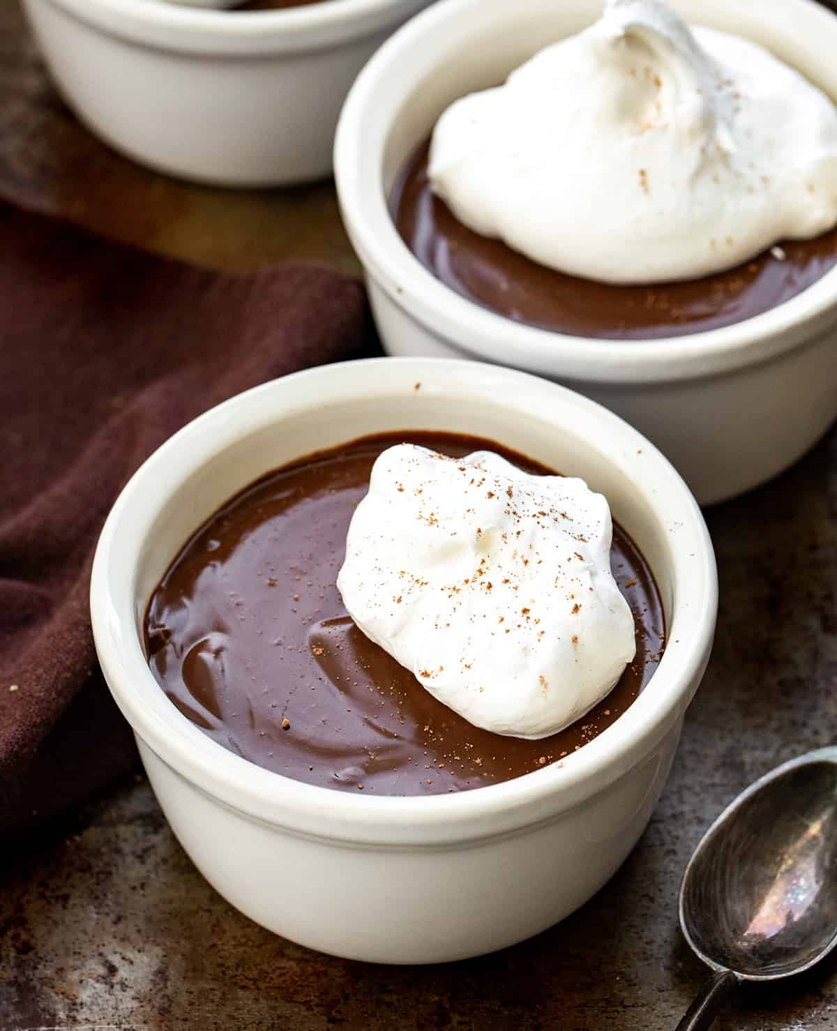 Indulge in Chocolate Pots de Crème, a creamy chocolate custard topped with whipped cream. Perfect for an elegant, easy homemade dessert! Chocolate Pots De Crème in white ramekins on a wooden table with a spoon.