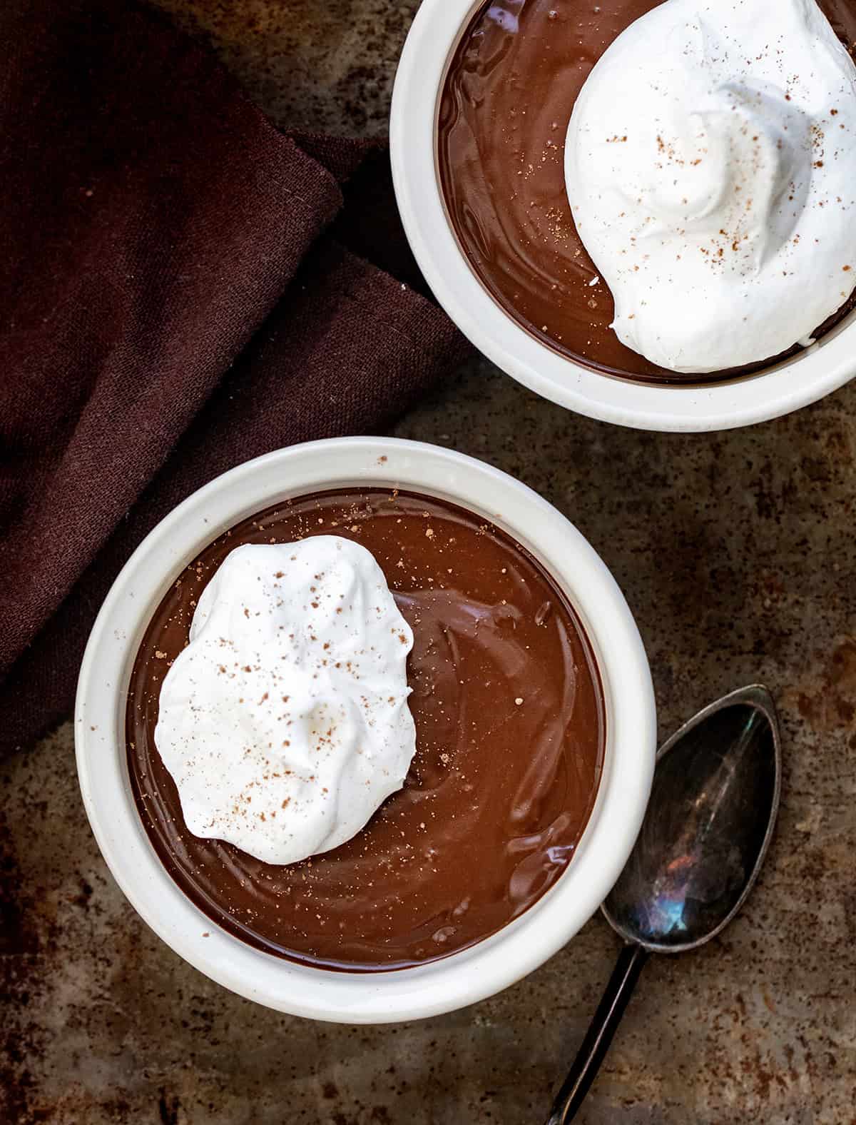 Indulge in Chocolate Pots de Crème, a creamy chocolate custard topped with whipped cream. Perfect for an elegant, easy homemade dessert! Chocolate Pots De Crème in white containers on a table from overhead.