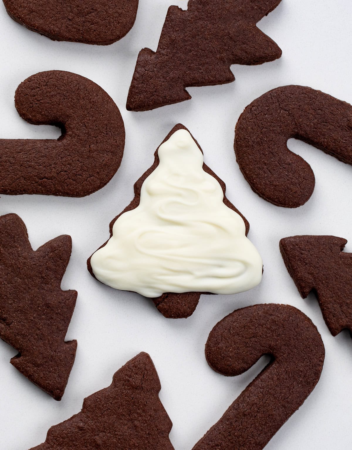 Cut-Out Chocolate Sugar Cookies are soft-centered, rich in cocoa, and hold their shape perfectly for decorating! Chocolate Sugar Cookies on a white table with the center cookie covered in white frosting.