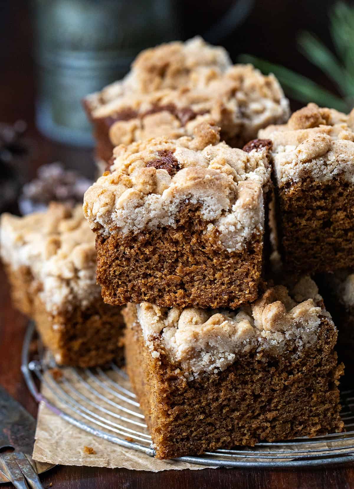 This Gingerbread Coffee Cake delivers warmth in every bite! The cake itself is soft, tender, and perfectly spiced with ginger, cinnamon, cloves, and nutmeg, sweetened with maple syrup and molasses! Pieces of Gingerbread Coffee Cake on a cool rack on a wooden table.