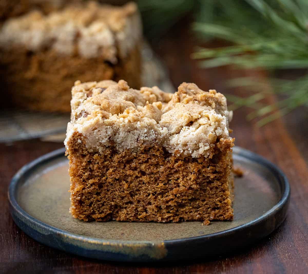 This Gingerbread Coffee Cake delivers warmth in every bite! The cake itself is soft, tender, and perfectly spiced with ginger, cinnamon, cloves, and nutmeg, sweetened with maple syrup and molasses! Piece of Gingerbread Coffee Cake on a small black plate on a wooden table.