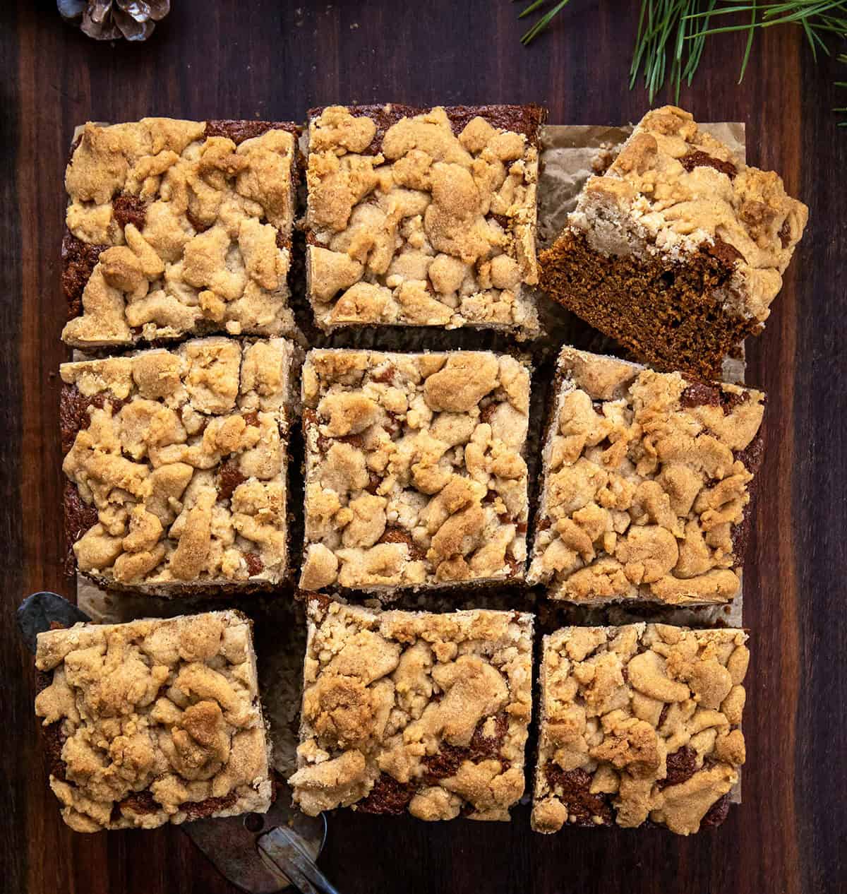 This Gingerbread Coffee Cake delivers warmth in every bite! The cake itself is soft, tender, and perfectly spiced with ginger, cinnamon, cloves, and nutmeg, sweetened with maple syrup and molasses! Gingerbread Coffee Cake cut into squares on a wooden table from overhead.