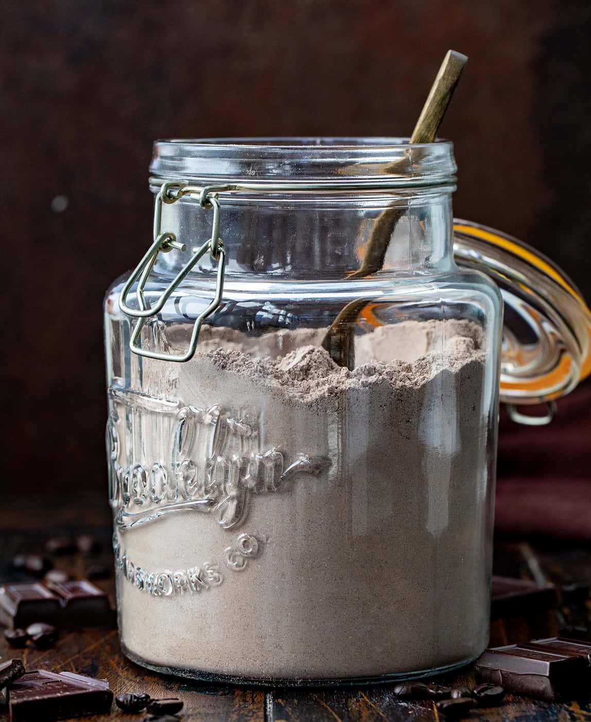 Warm up your mornings with this Mocha Hot Cocoa Mix! Rich cocoa, a hint of espresso, and sweet powdered milk come together to create a creamy, chocolatey mug of comfort in minutes! Jar of Mocha Hot Cocoa Mix with a spoon in it on a wooden table.