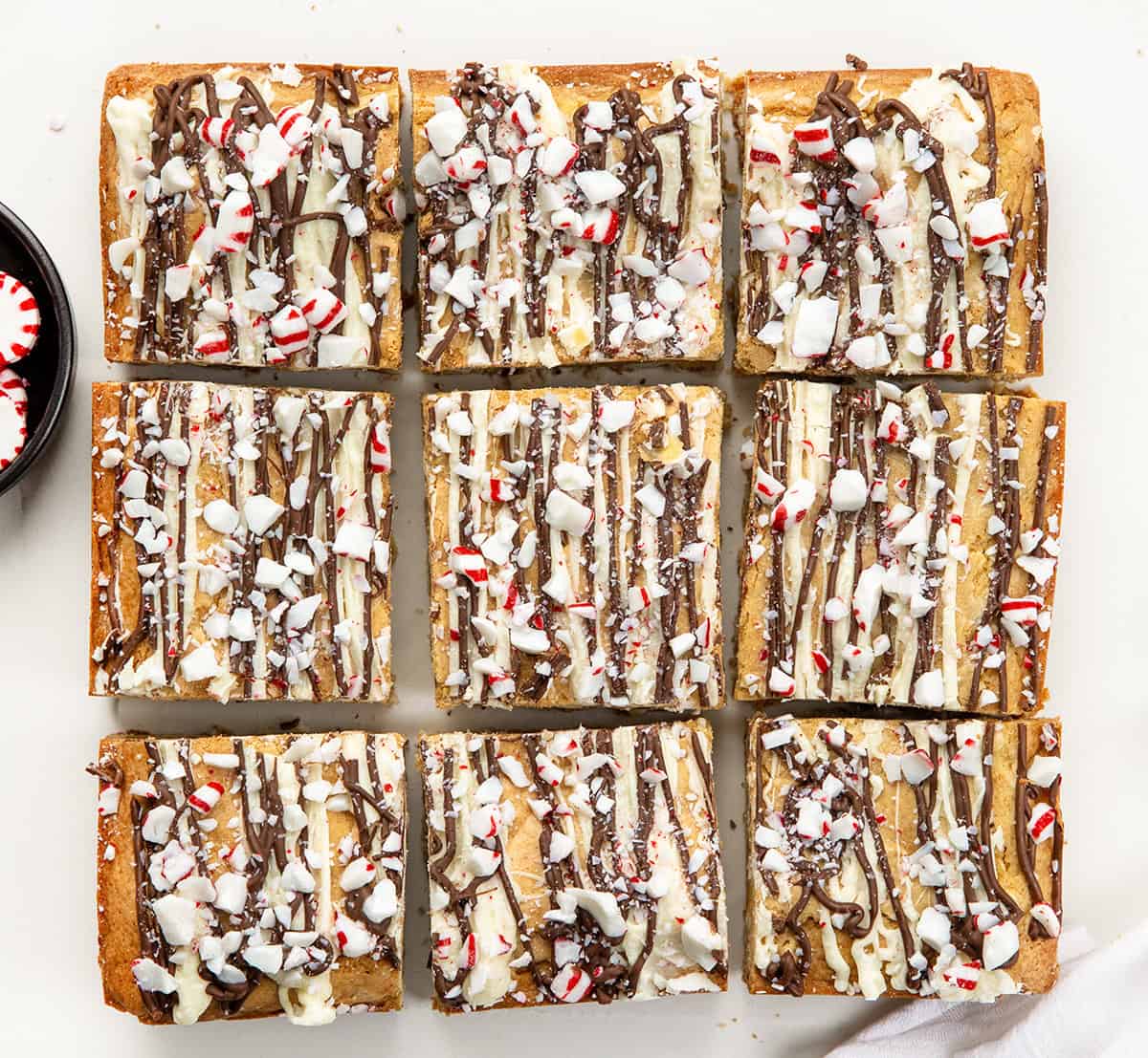 These Peppermint Bark Blondies are rich, buttery, and festive, the perfect holiday treat! The base is my chewy white chocolate blondie, flavored with a touch of peppermint extract. I top each blondie with a double drizzle of milk and white chocolate, then sprinkle on crushed peppermint candies for a holiday-perfect crunch! Peppermint Bark Blondies cut into squares on a white table from overhead.