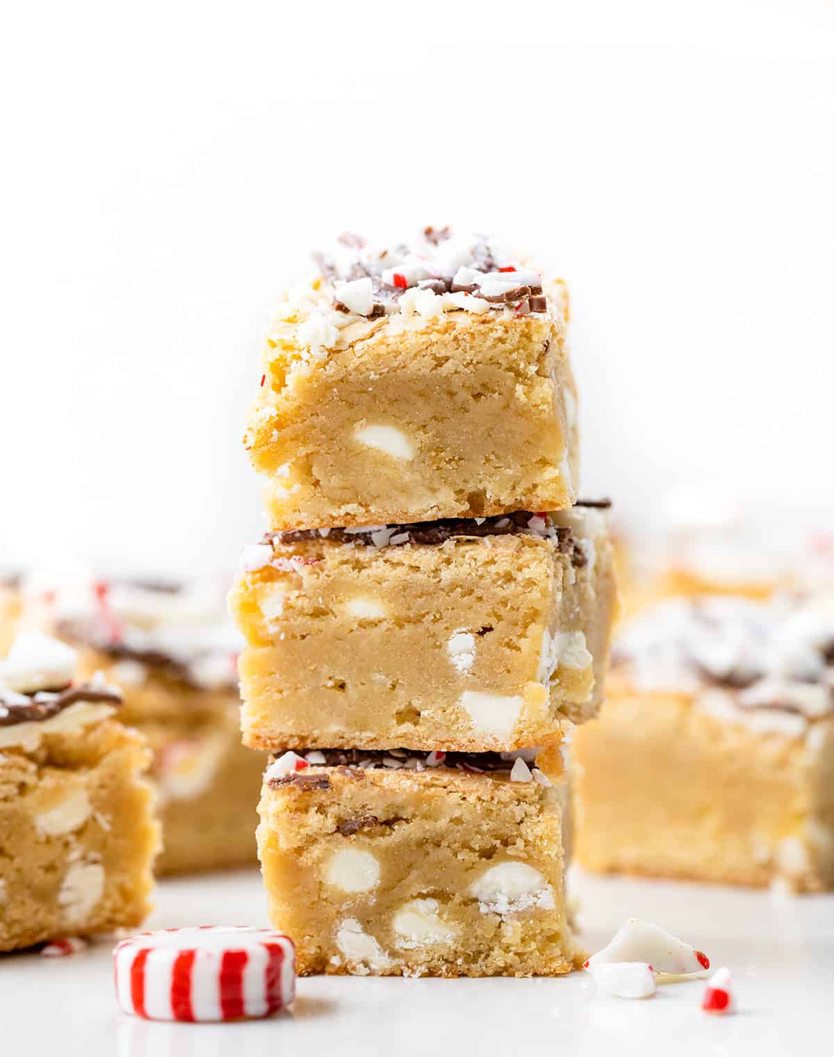 These Peppermint Bark Blondies are rich, buttery, and festive, the perfect holiday treat! The base is my chewy white chocolate blondie, flavored with a touch of peppermint extract. I top each blondie with a double drizzle of milk and white chocolate, then sprinkle on crushed peppermint candies for a holiday-perfect crunch! Stack of Peppermint Bark Blondies on a white table.