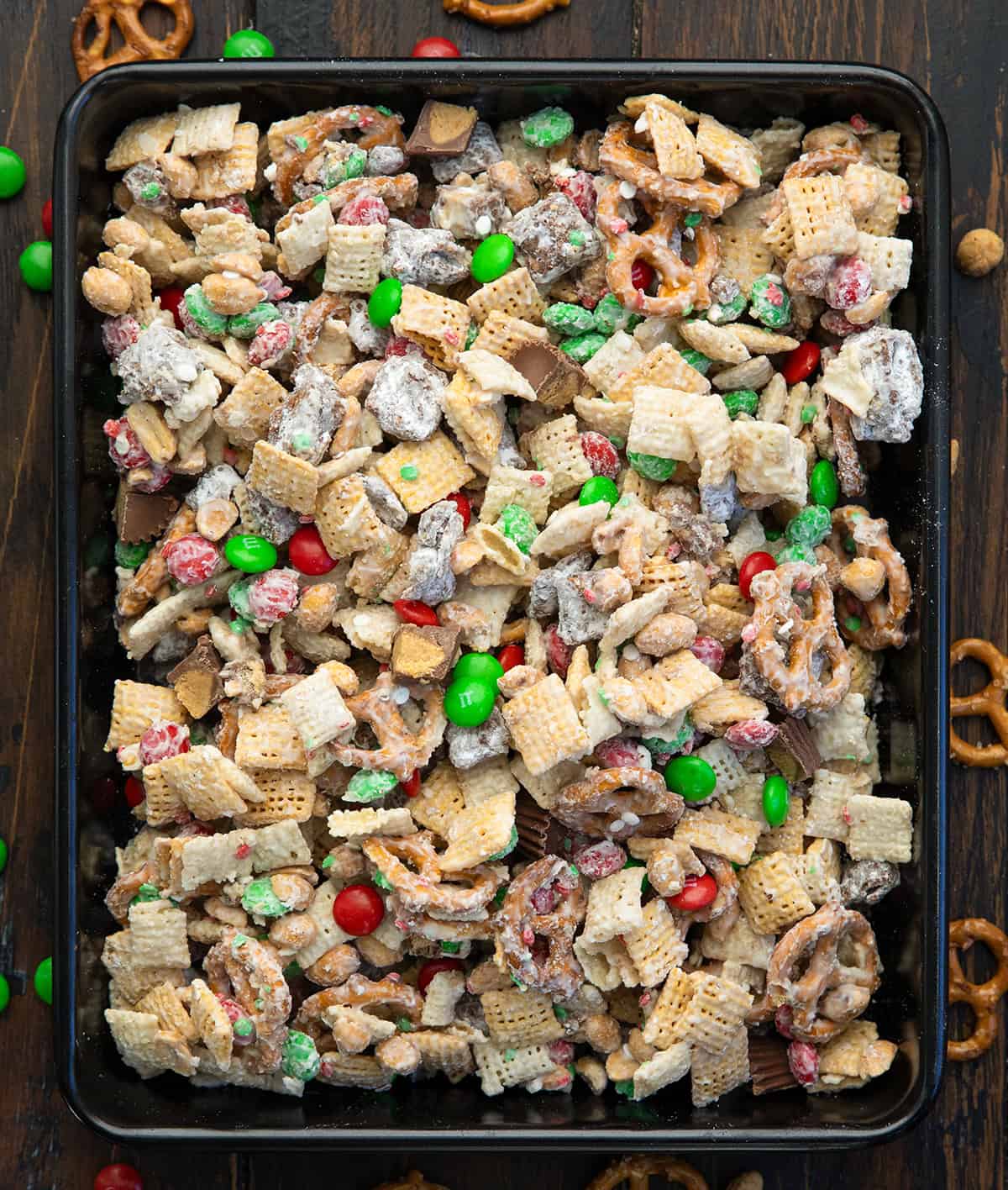 This Ultimate Christmas Snack Mix is what happens when all of my favorite holiday treats collide into one bowl. It’s sweet, salty, crunchy, and truly over-the-top... combining classic peanut butter puppy chow, glossy white chocolate-coated Chex, salty pretzels, peanuts, and festive holiday candies! Black tray of Ultimate Christmas Snack Mix on a wooden table from overhead.