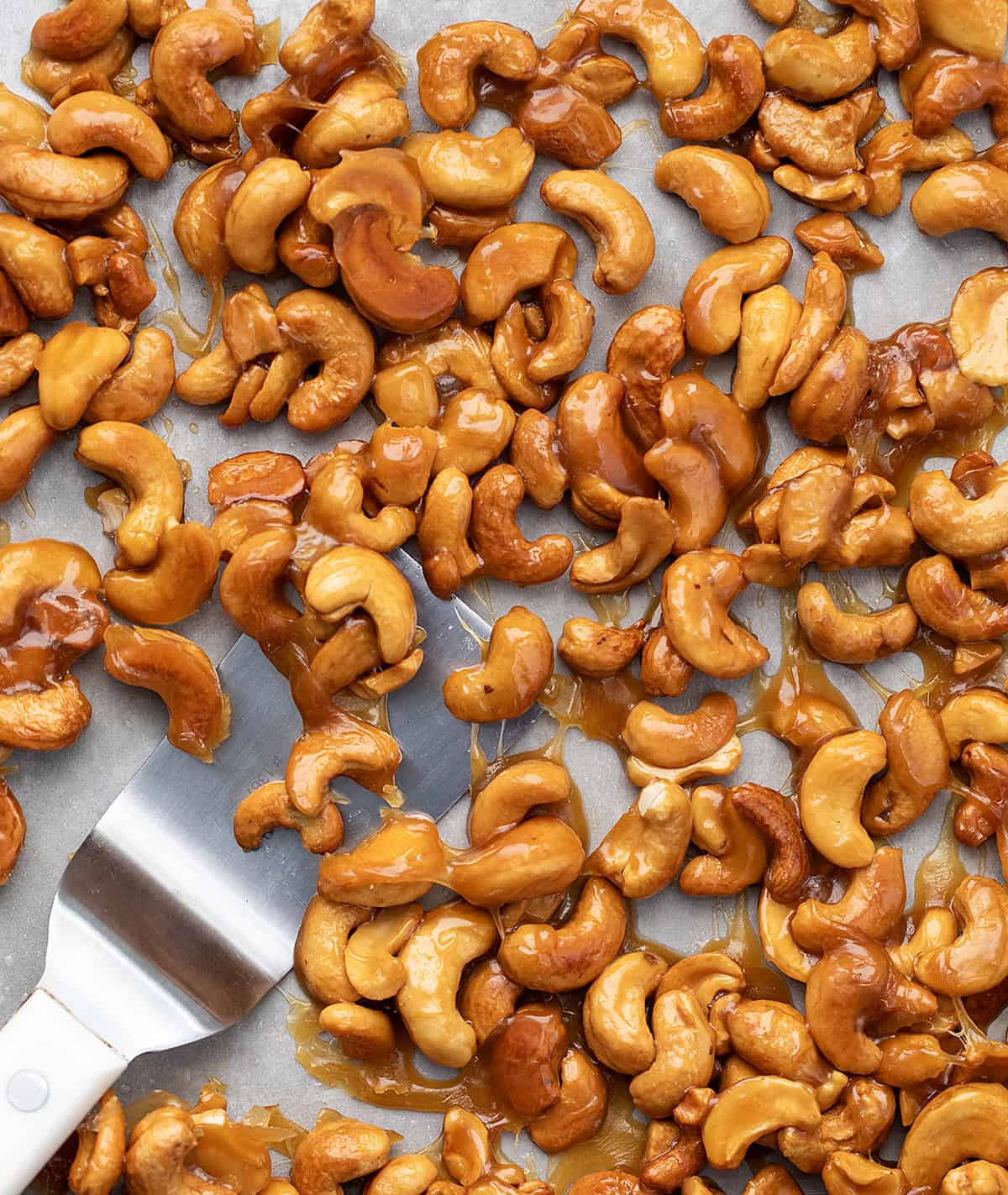 Candied Cashews are sweet, crunchy, and totally addictive. A quick toss in honey, brown sugar, and butter turns raw cashews into a golden, caramel-y snack that’s perfectly delicious! Close up of Candied Cashews on a pan with a spatula.