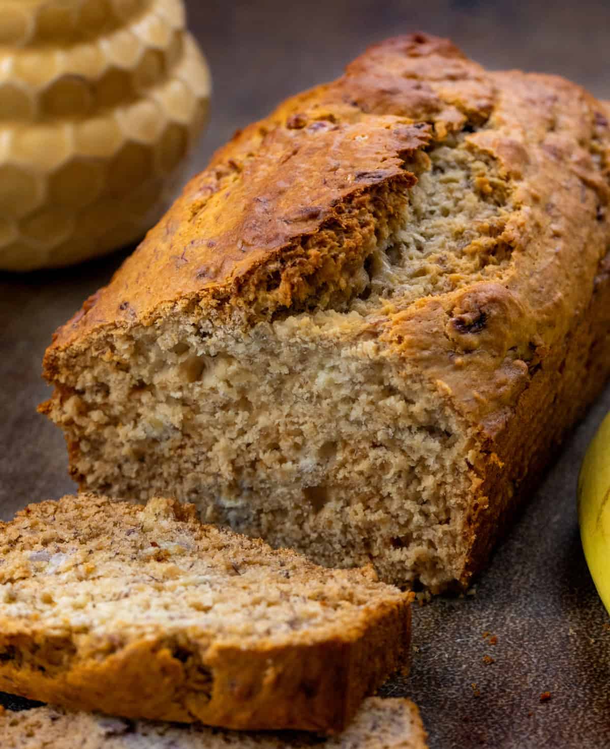 This Honey Banana Bread is a cozy quick bread that’s naturally sweetened with honey instead of sugar. The bananas keep the loaf soft and moist, and the honey gives a warm flavor and golden edges! Loaf of Honey Banana Bread that has a slice cut off and laying flat with a honey pot and banana.