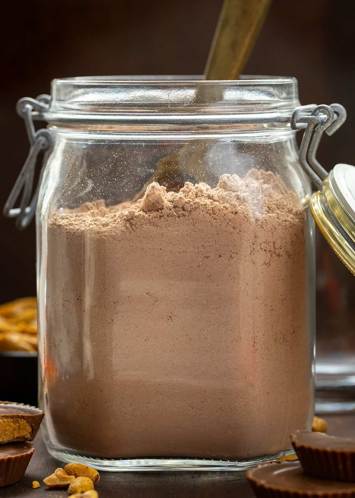 If you love the cozy comfort of homemade hot cocoa but want something a little richer and more fun, this Peanut Butter Hot Cocoa Mix is going to be your new winter favorite. It blends smooth peanut butter flavor with deep, chocolatey warmth! Container of Peanut Butter Hot Cocoa Mix on a wooden table with reese's peanut butter cups and peanut butter around.