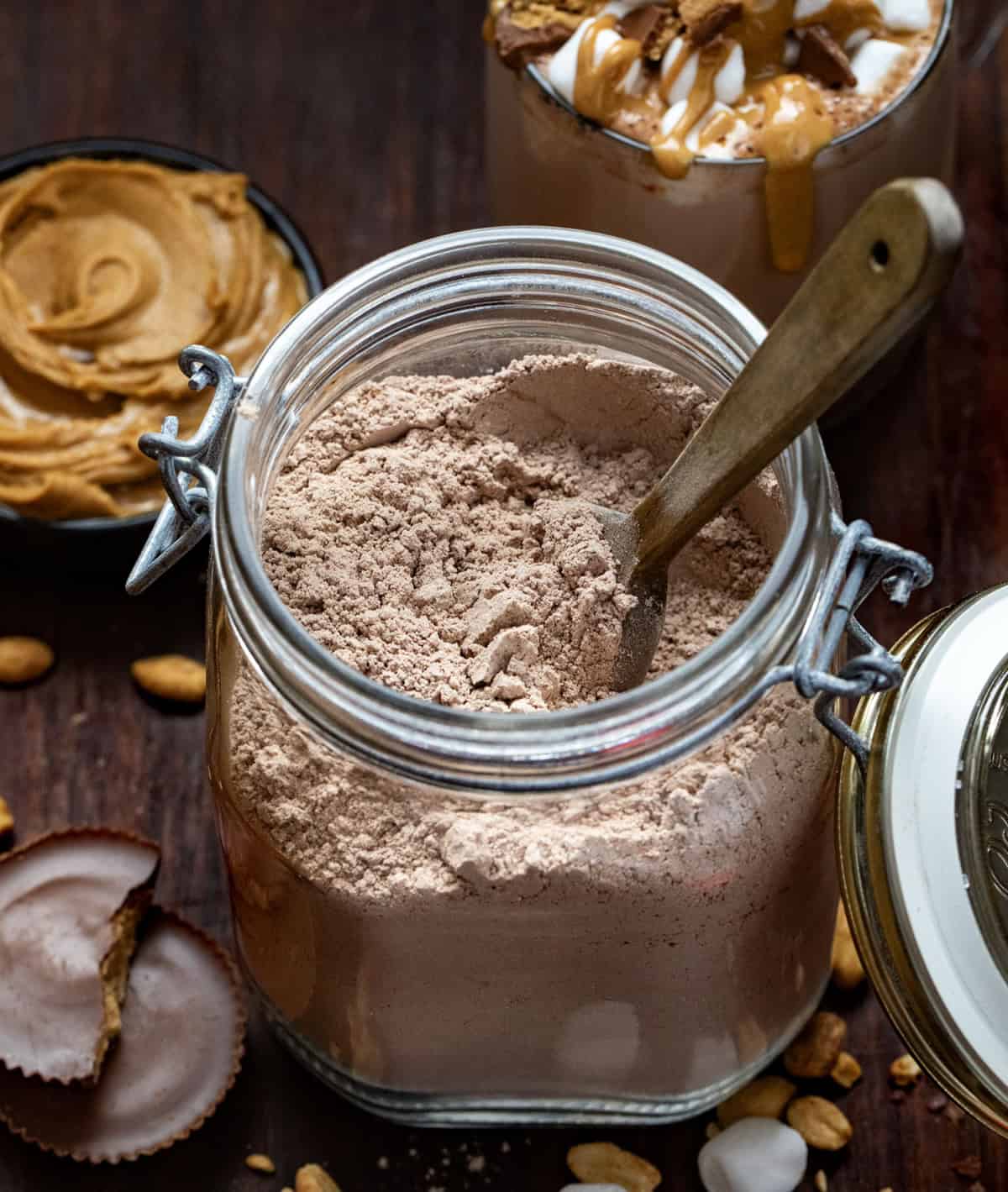 Jar of Peanut Butter Hot Cocoa Mix with a wooden spoon in it with peanut butter in the background.