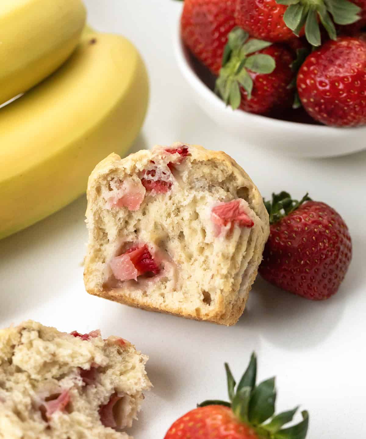 Halved Strawberry Banana Muffin on a white table with bananas and fresh strawberries.