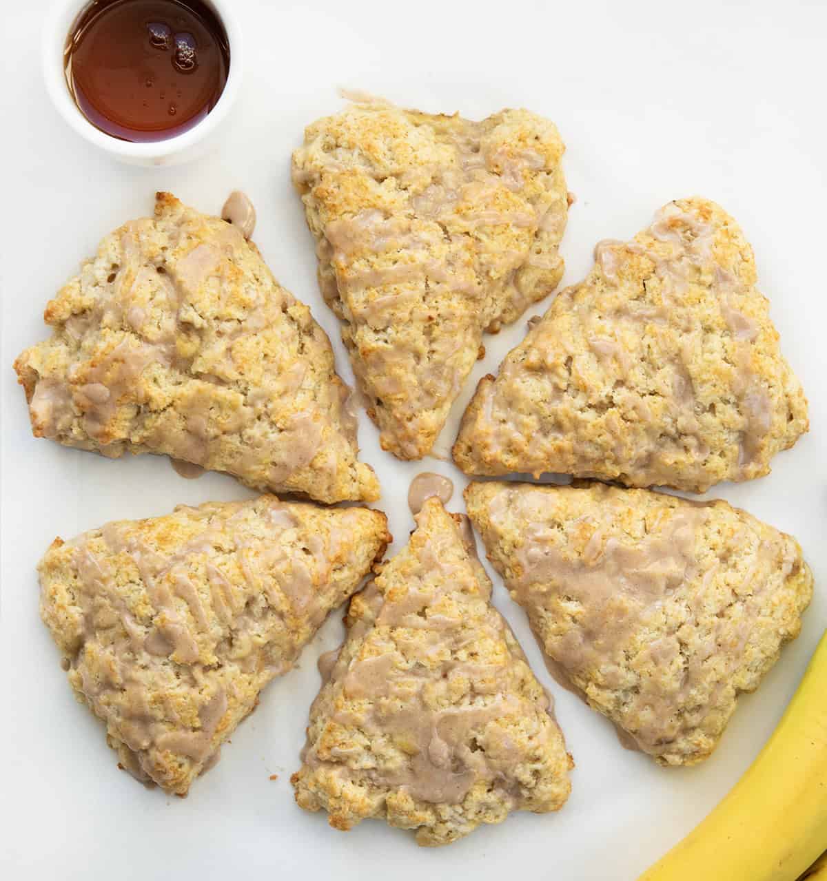 I love these banana scones with maple glaze because they feel like something from a cute bakery case! The edges bake up crisp and golden, and the centers stay soft and tender. The cinnamon maple glaze on top adds sweetness and makes them feel extra special! Banana Scones laid out in a circle with maple syrup and bananas.
