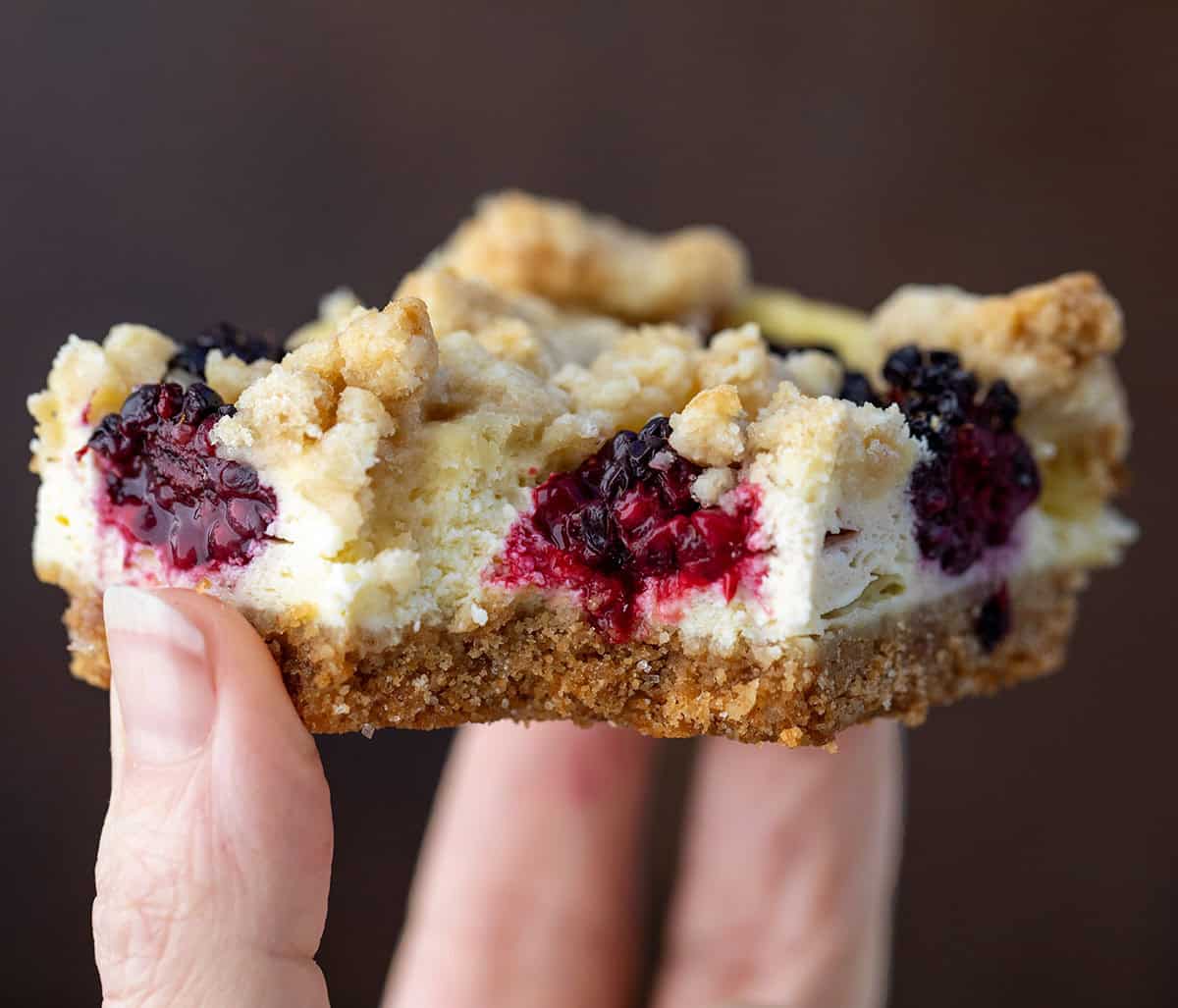 Blackberry Cheesecake Bars are creamy, fruity, and so easy to slice and share. Fresh blackberries add bright pops of flavor to the smooth cheesecake filling and a buttery graham crust that makes every bite irresistible! Hand holding a bit into Blackberry Cheesecake Bar.