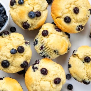 Blueberry Buttermilk Muffins on a white table with fresh blueberries and some muffins on their side.