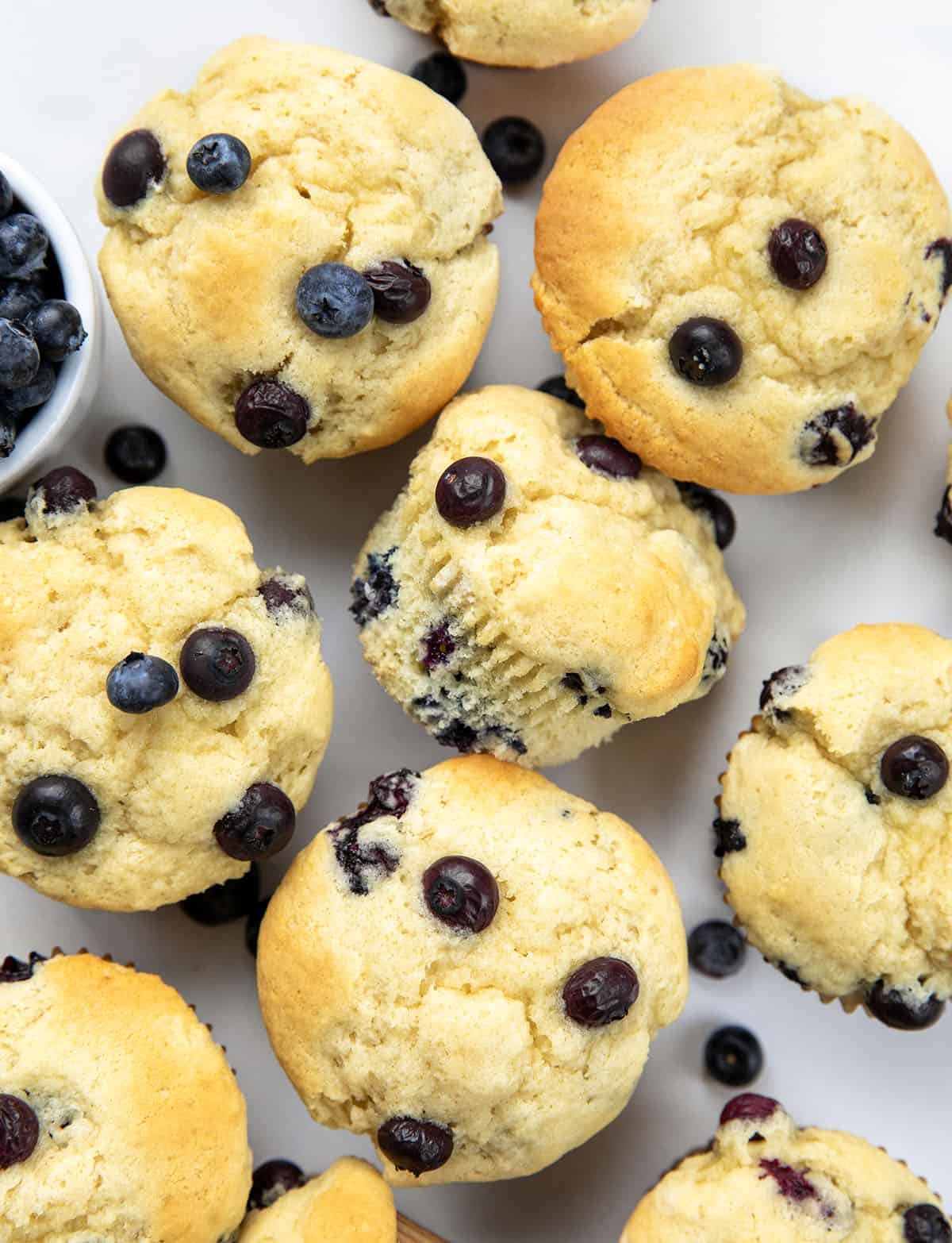 Blueberry Buttermilk Muffins on a white table with fresh blueberries and some muffins on their side.