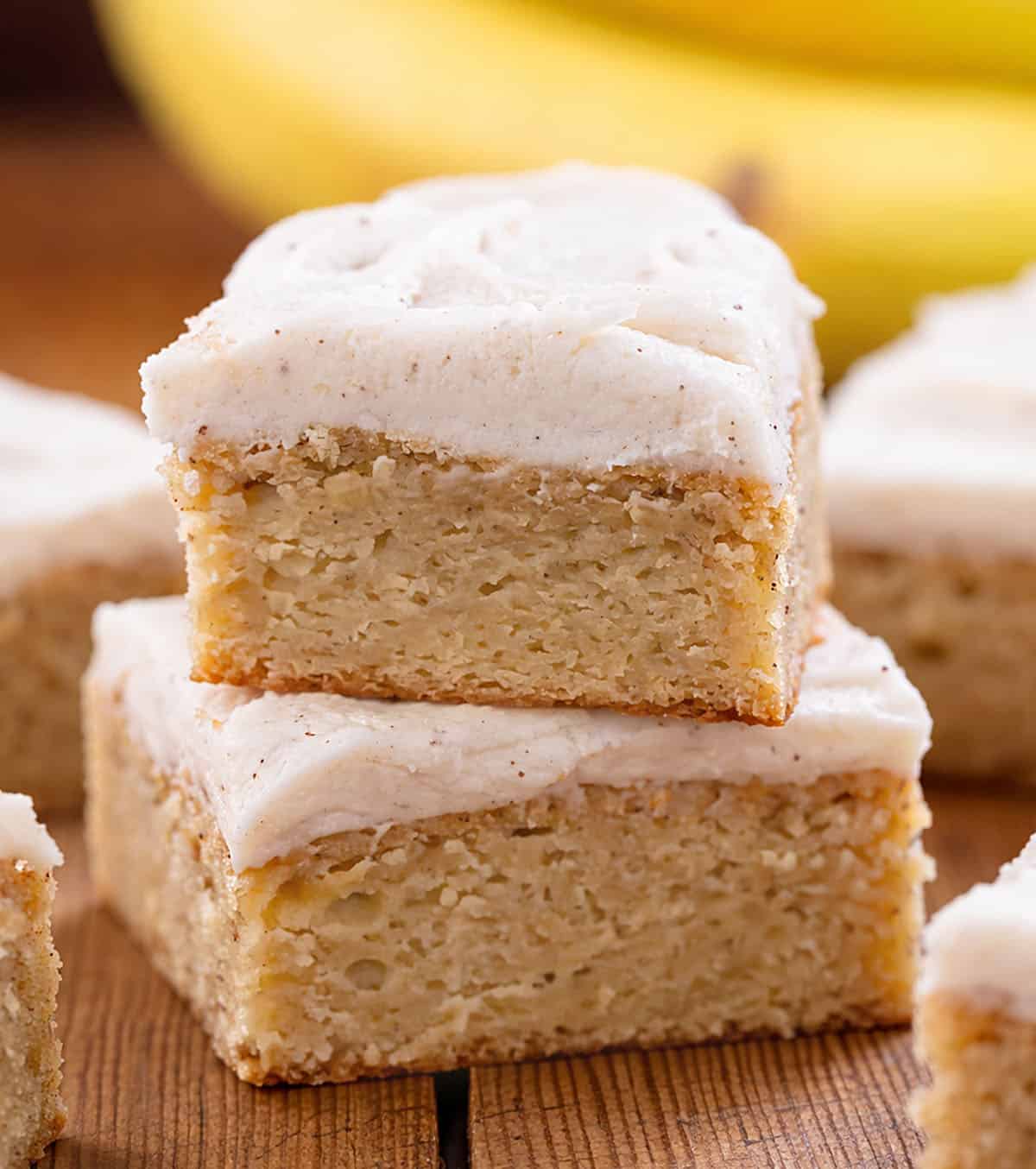 Stack of Browned Butter Banana Bars with Browned Butter Frosting on a wooden table with bananas in the background.