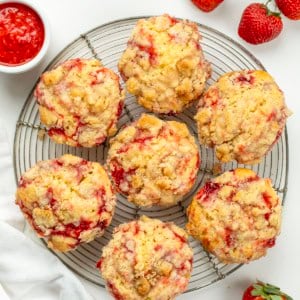 Double Strawberry Muffins on a round wire rack on a white table with strawberry sauce and fresh strawberries.