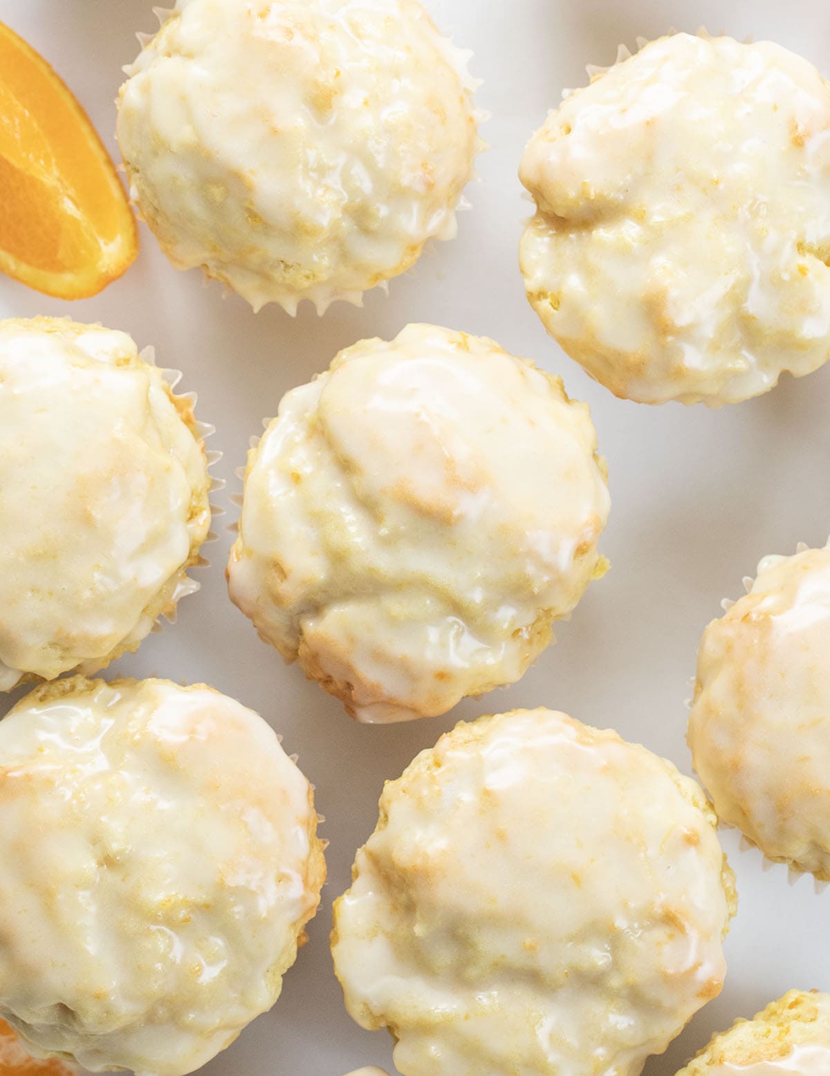 Bright, fluffy, and bursting with fresh orange flavor, these orange vanilla buttermilk muffins are a bakery-style treat you can make right at home. The combination of citrus zest, warm vanilla, and tender buttermilk crumb makes every bite soft, citrusy, and completely irresistible! Glazed Orange Vanilla Buttermilk Muffins on a white table with an orange slice from overhead.