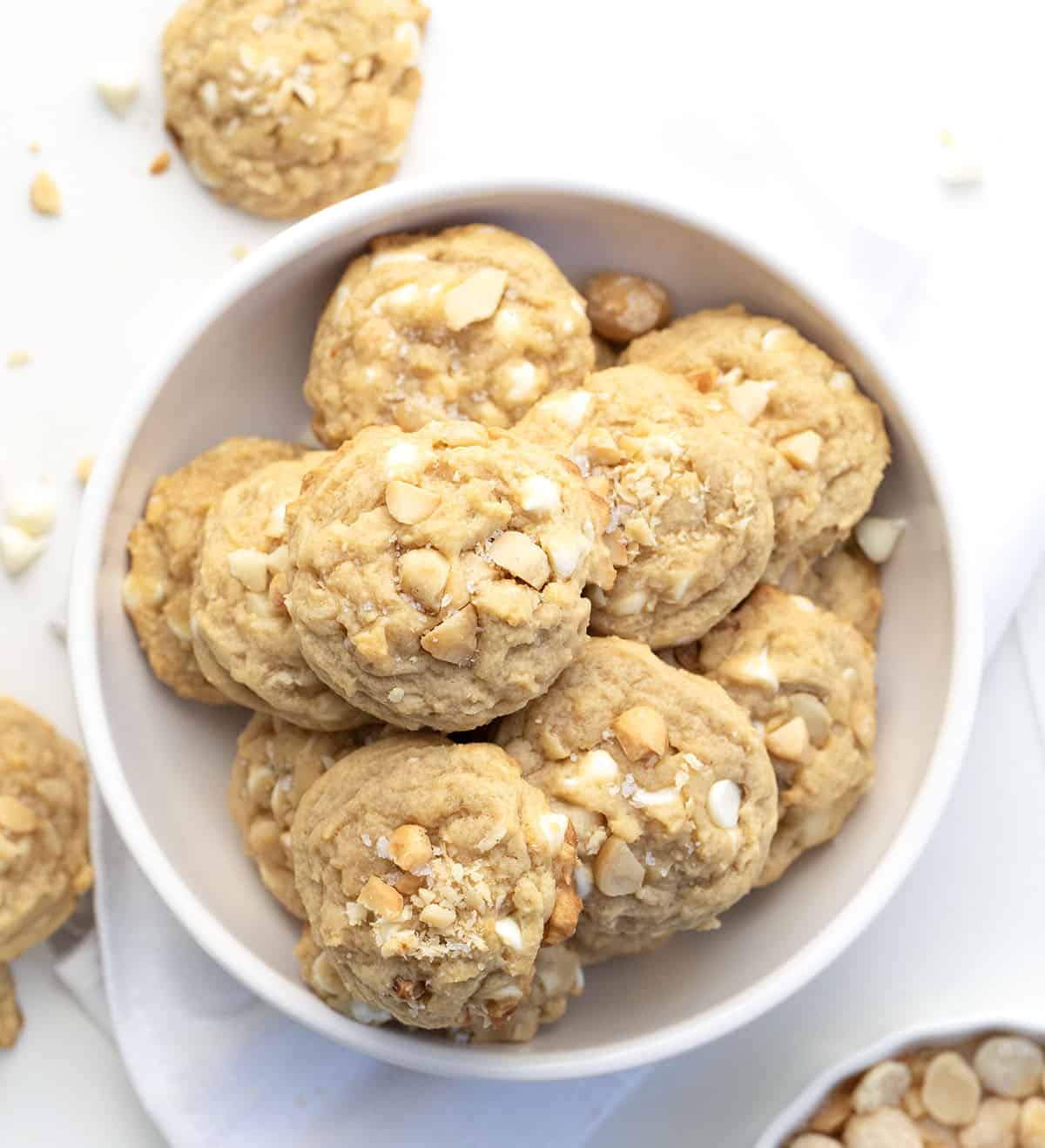 Golden, buttery, and packed with white chocolate and macadamia nuts! These Salted Honey White Chocolate Macadamia Nut Cookies are soft in the center, lightly crisp at the edges, and sprinkled with just the right touch of flaky sea salt! White bowl filled with Salted Honey White Chocolate Macadamia Nut Cookies on a white table from overhead.