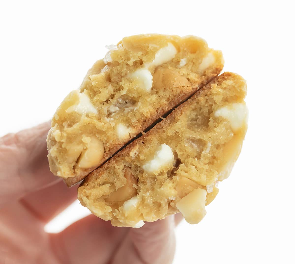 Golden, buttery, and packed with white chocolate and macadamia nuts! These Salted Honey White Chocolate Macadamia Nut Cookies are soft in the center, lightly crisp at the edges, and sprinkled with just the right touch of flaky sea salt! Hand holding a halved Salted Honey White Chocolate Macadamia Nut Cookie.