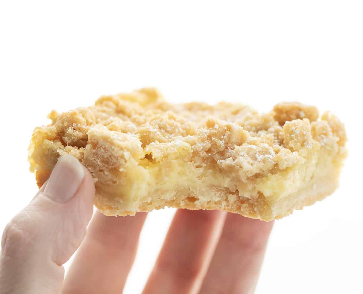 These Vanilla Cream Crumb Bars start with a buttery shortbread crust layered with a smooth vanilla custard filling, and are finished with a golden brown crumble topping! Hand holding a bit into Vanilla Cream Crumb Bar.
