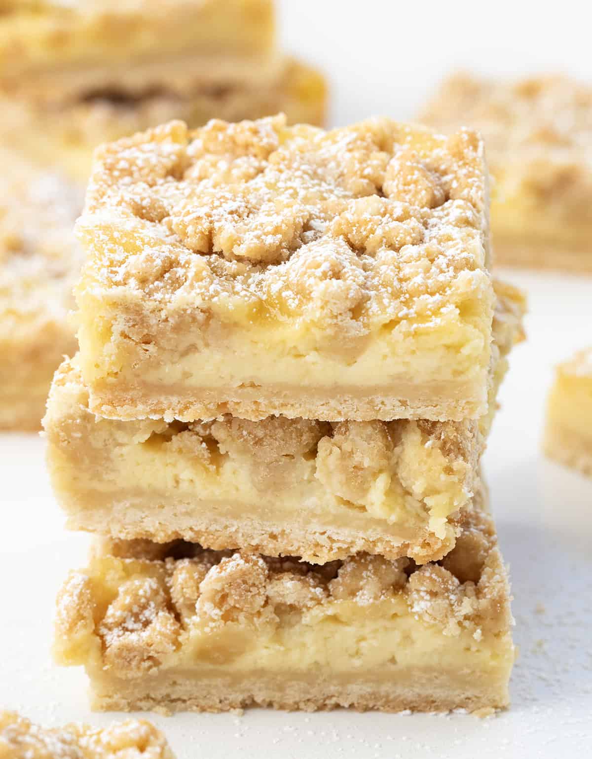 These Vanilla Cream Crumb Bars start with a buttery shortbread crust layered with a smooth vanilla custard filling, and are finished with a golden brown crumble topping! Stack of Vanilla Cream Crumb Bars on a white table surrounded by other bars.