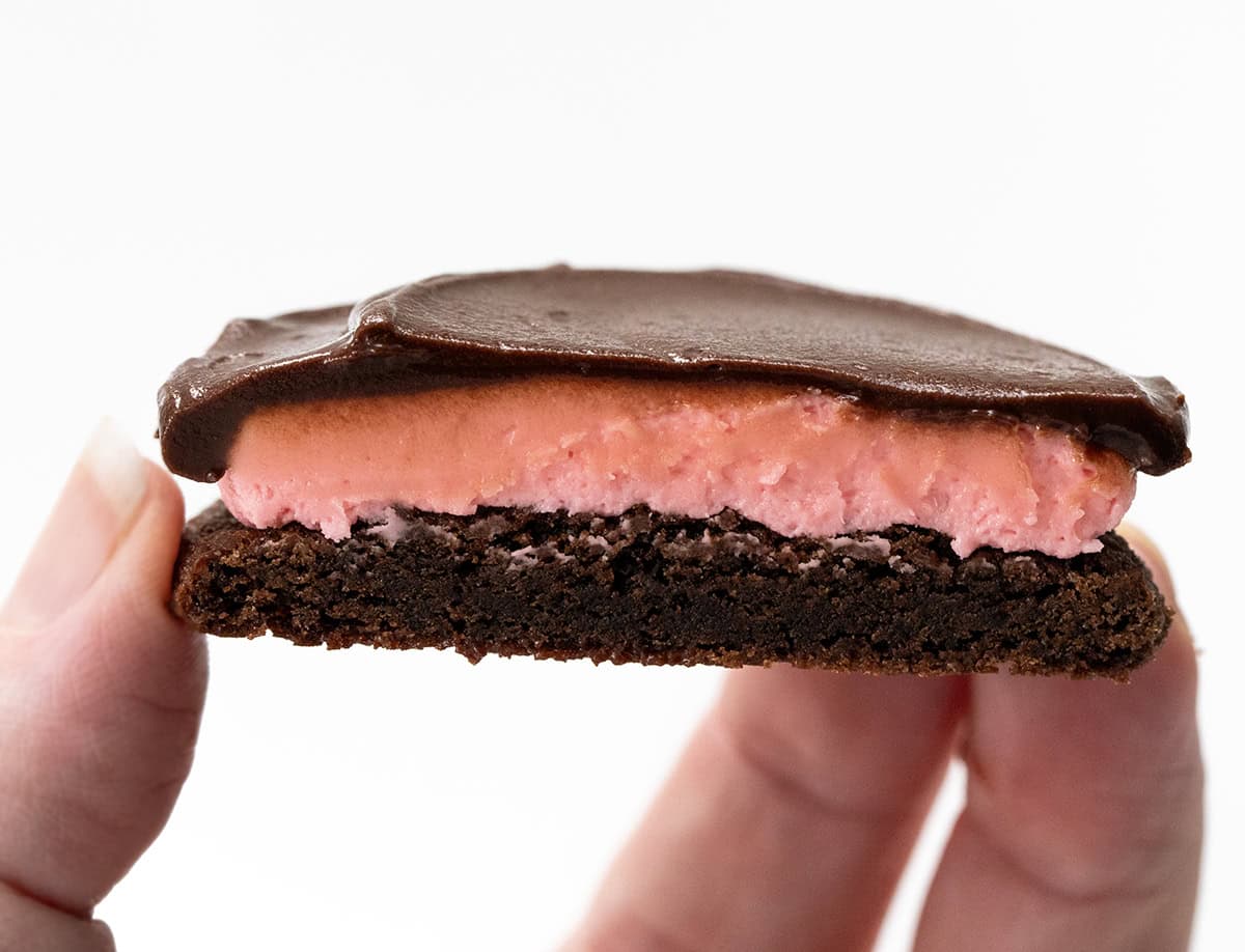 Soft, chocolatey cookies topped with a sweet and tangy raspberry buttercream and finished with a shiny chocolate ganache. They are rich, chewy, and absolutely delicious! Hand holding a halved Chocolate Raspberry Cookie showing the layers.