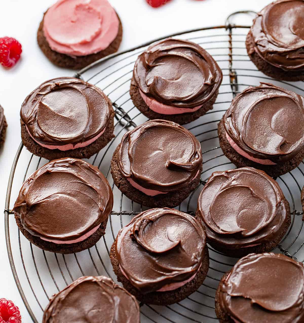 Soft, chocolatey cookies topped with a sweet and tangy raspberry buttercream and finished with a shiny chocolate ganache. They are rich, chewy, and absolutely delicious! Chocolate Raspberry Cookies on a cooling rack on a white table with fresh raspberries.