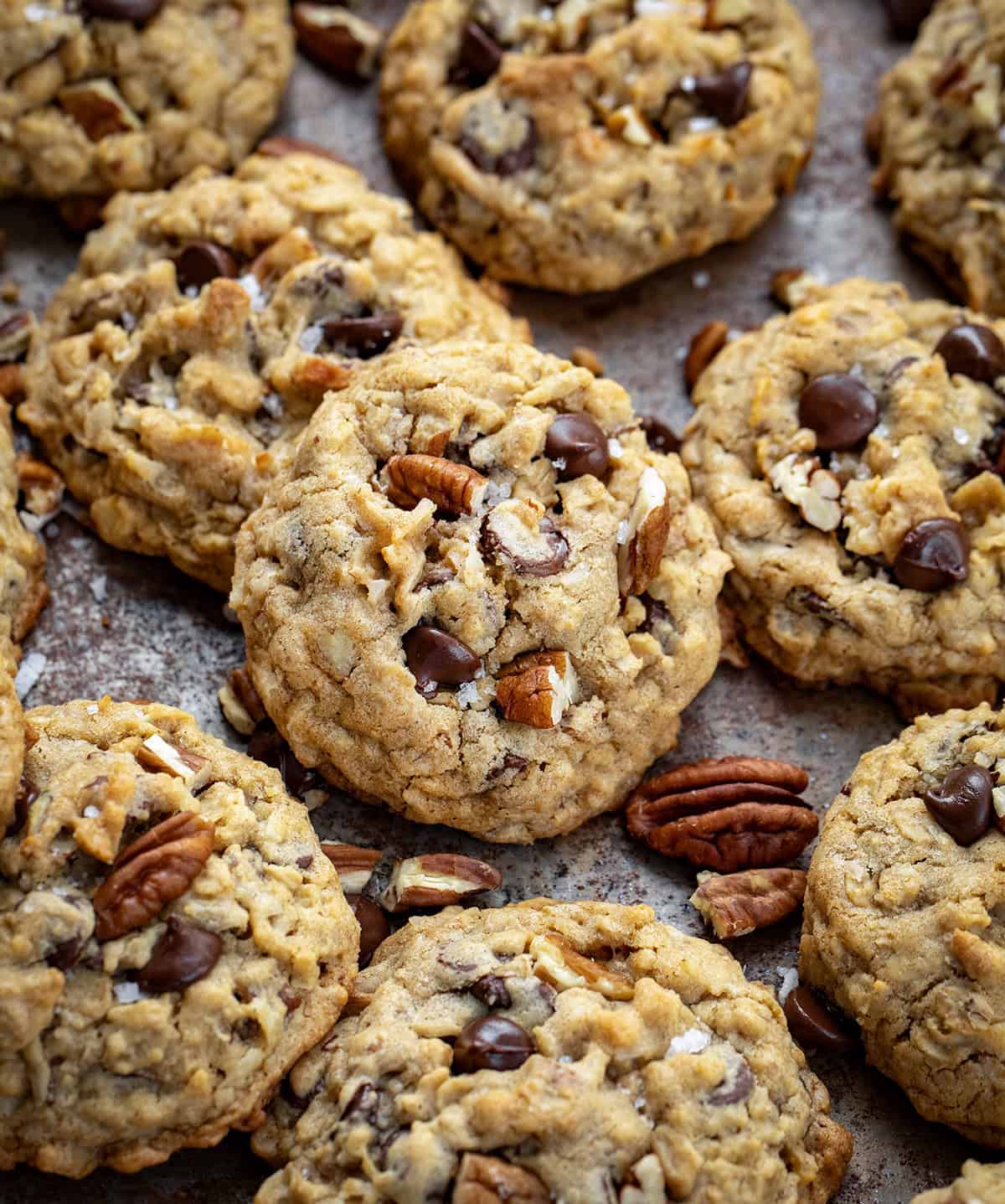 These Cowboy Cookies are thick, chewy, and loaded with oats, chocolate chips, pecans, and coconut. Each bite is soft in the middle with lightly golden edges and a little crunch from the nuts and coconut! Cowboy Cookies on a dark sheet pan with pecans and coconut flakes.