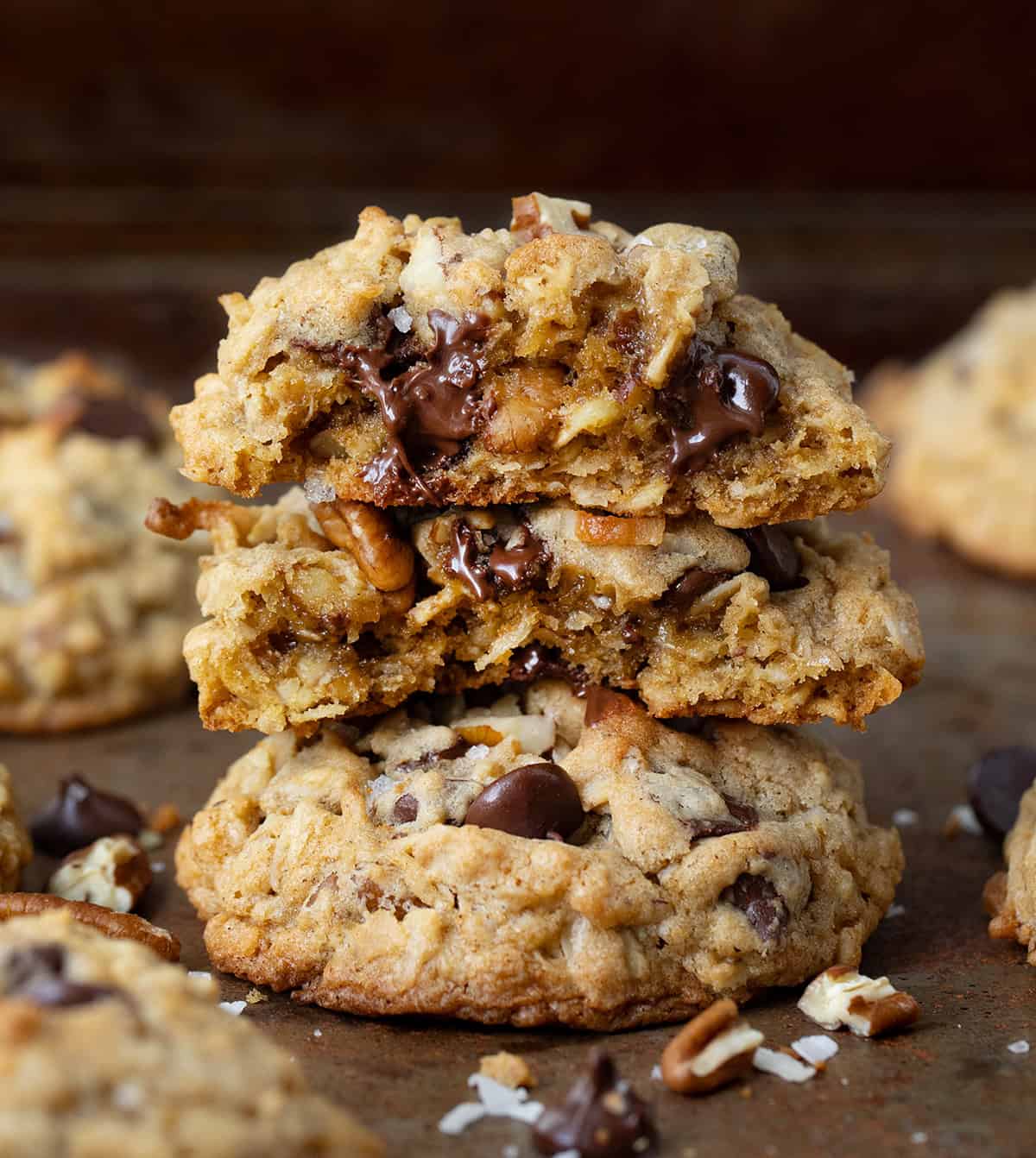 These Cowboy Cookies are thick, chewy, and loaded with oats, chocolate chips, pecans, and coconut. Each bite is soft in the middle with lightly golden edges and a little crunch from the nuts and coconut! Stacked Cowboy Cookies on a sheet pan with top cookie halved showing inside texture.
