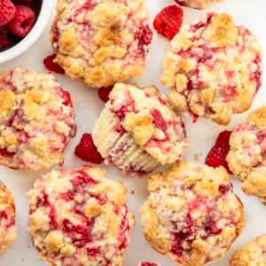 Looking down on a white table covered with Double Raspberry Muffins, with one muffin on its side and fresh raspberries.