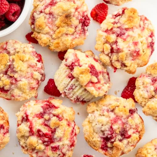 Looking down on a white table covered with Double Raspberry Muffins, with one muffin on its side and fresh raspberries.