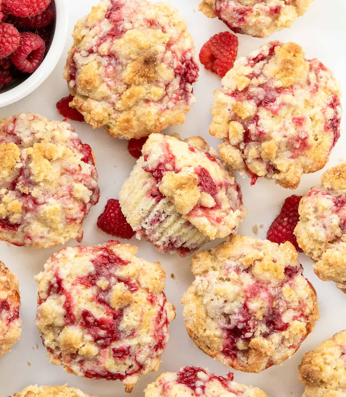 Looking down on a white table covered with Double Raspberry Muffins, with one muffin on its side and fresh raspberries.