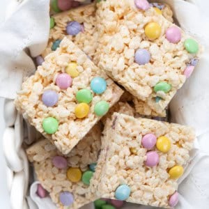 White glass basket of Easter Rice Krispie Treats cut into squares and stacked on a white napkin.