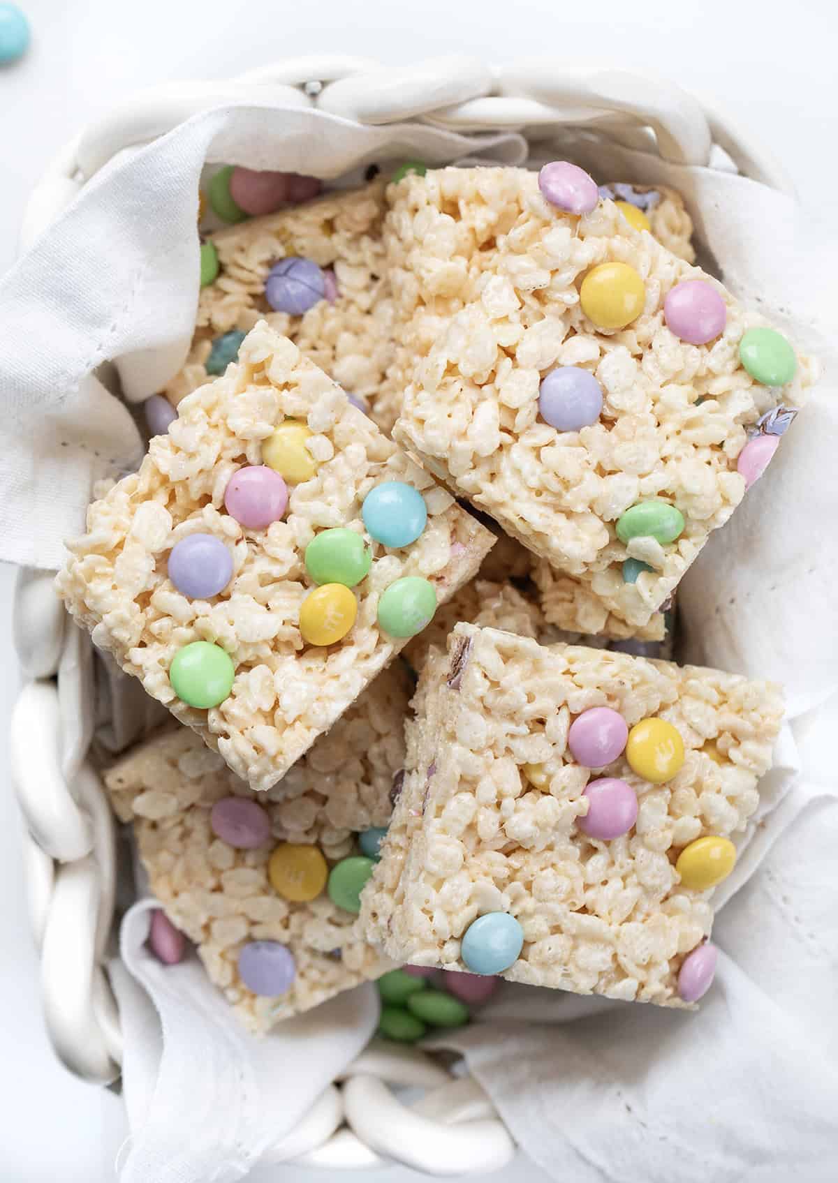 White glass basket of Easter Rice Krispie Treats cut into squares and stacked on a white napkin.