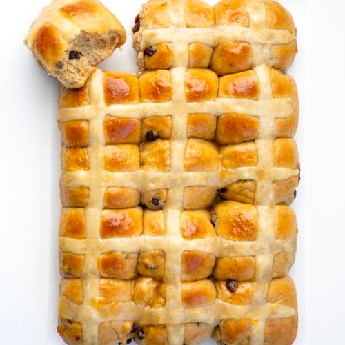 Hot Cross Buns on a white table with one bun pulled apart from the rest and on its side.