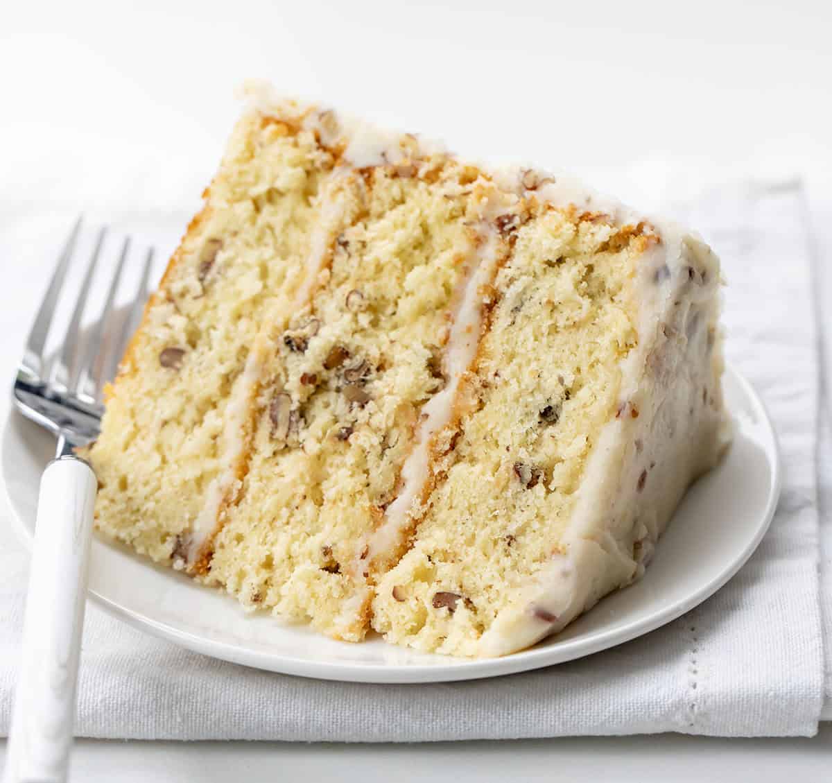 White plate with a piece of Italian Cream Cake on it with a white fork resting on the plate.