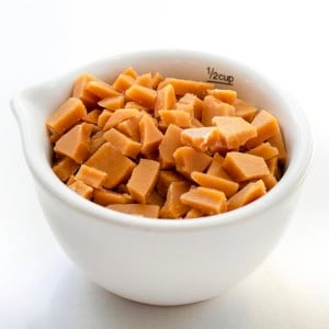 White measuring cup filled with Toffee Bits on a white table close up.