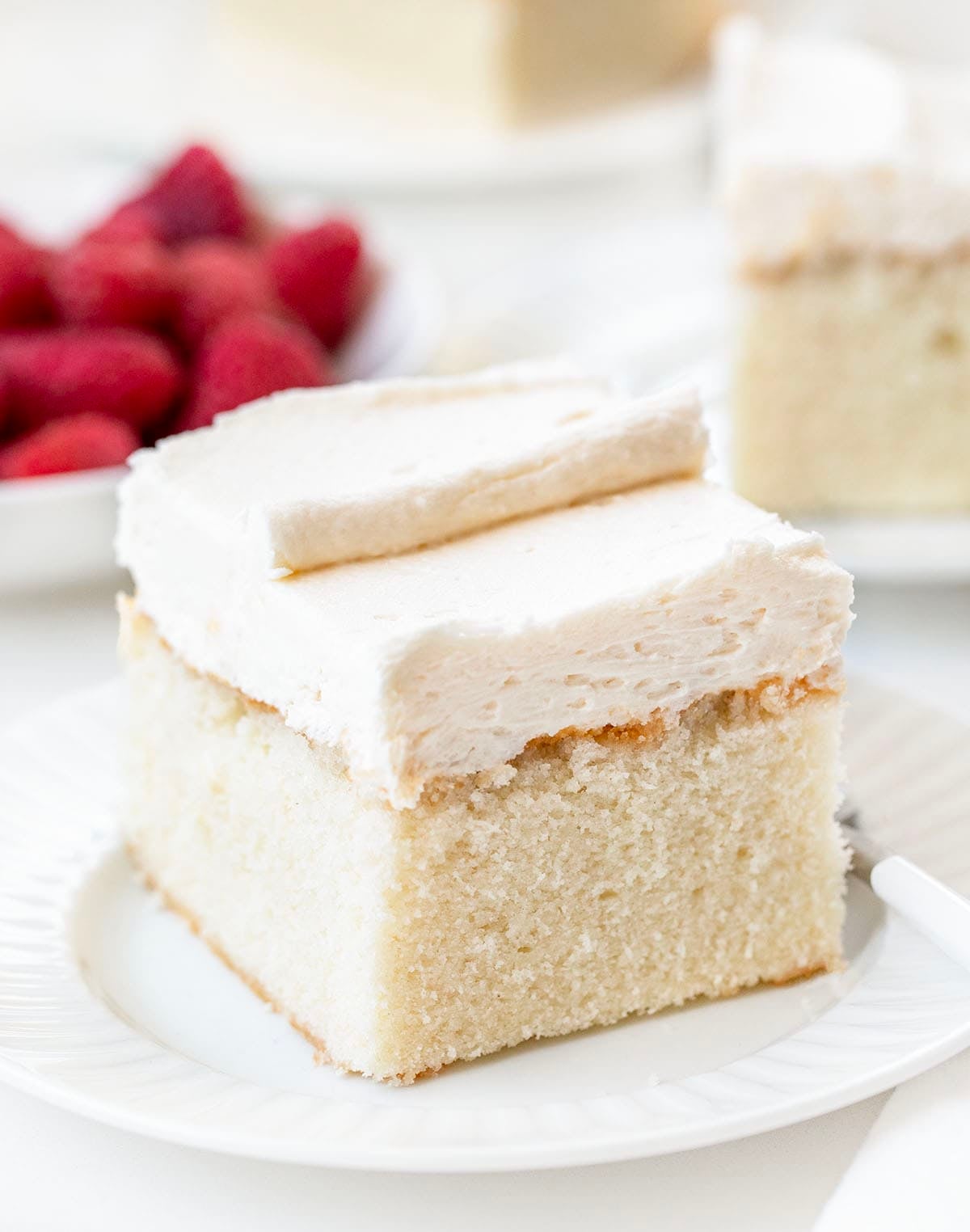 Piece of Best White Sheet Cake on a white plate on a white table with fresh raspberries.