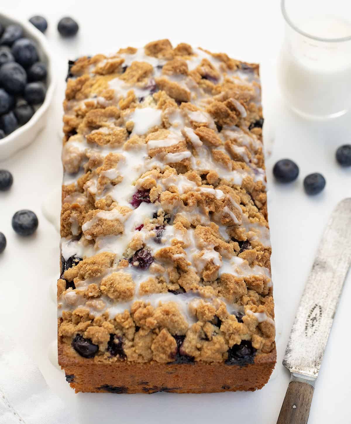 Loaf of Blueberry Crumble Bread on a white table with blueberries, glaze, and a knife.