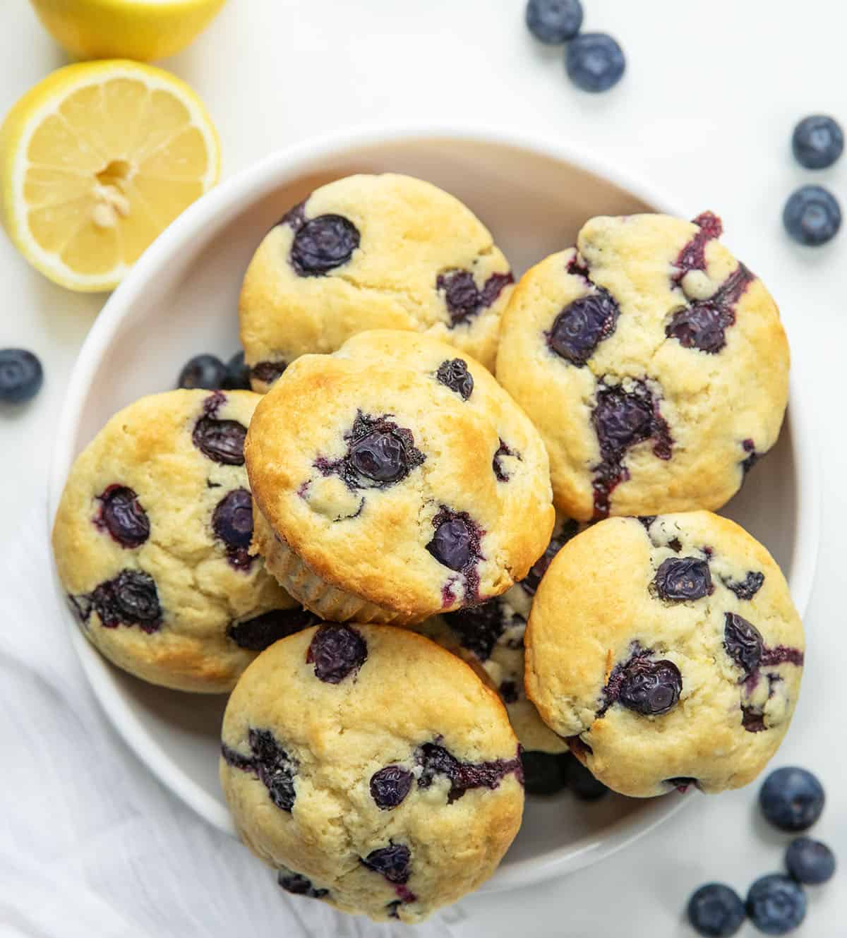 Bowl of Blueberry Lemon Muffin on a white table with blueberries and lemons from overhead.
