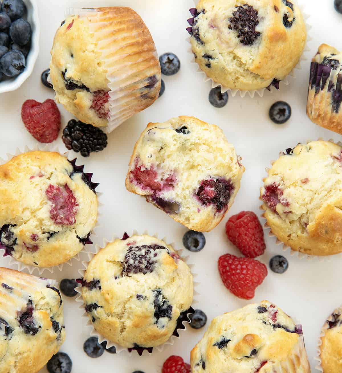 Soft, tender, and filled with a triple threat of flavor, these Triple Berry Muffins are packed with juicy blueberries, raspberries, and blackberries in every bite! Triple Berry Muffins on a white table with the center muffin halved from overhead.