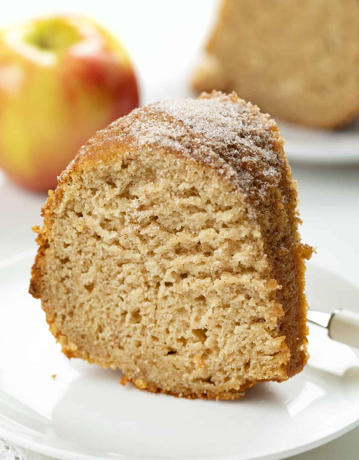 Slice of Apple Cider Donut Bundt Cake on a white plate with an apple in the background.