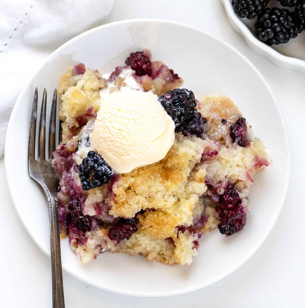 White plate with a portion of Blackberry Cobbler and ice cream on it with a fork resting on the side.
