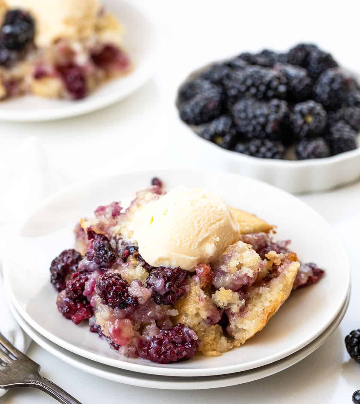 Plates of Blackberry Cobbler and a bowl of blackberries on a white table.
